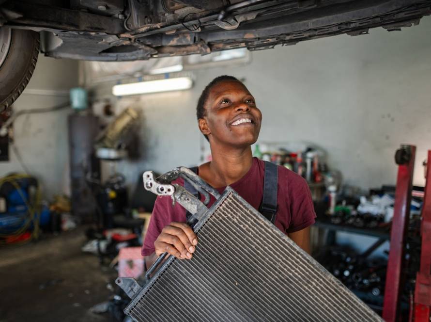 Female auto mechanic holding a radiator while inspecting the car underneath in a professional repair shop