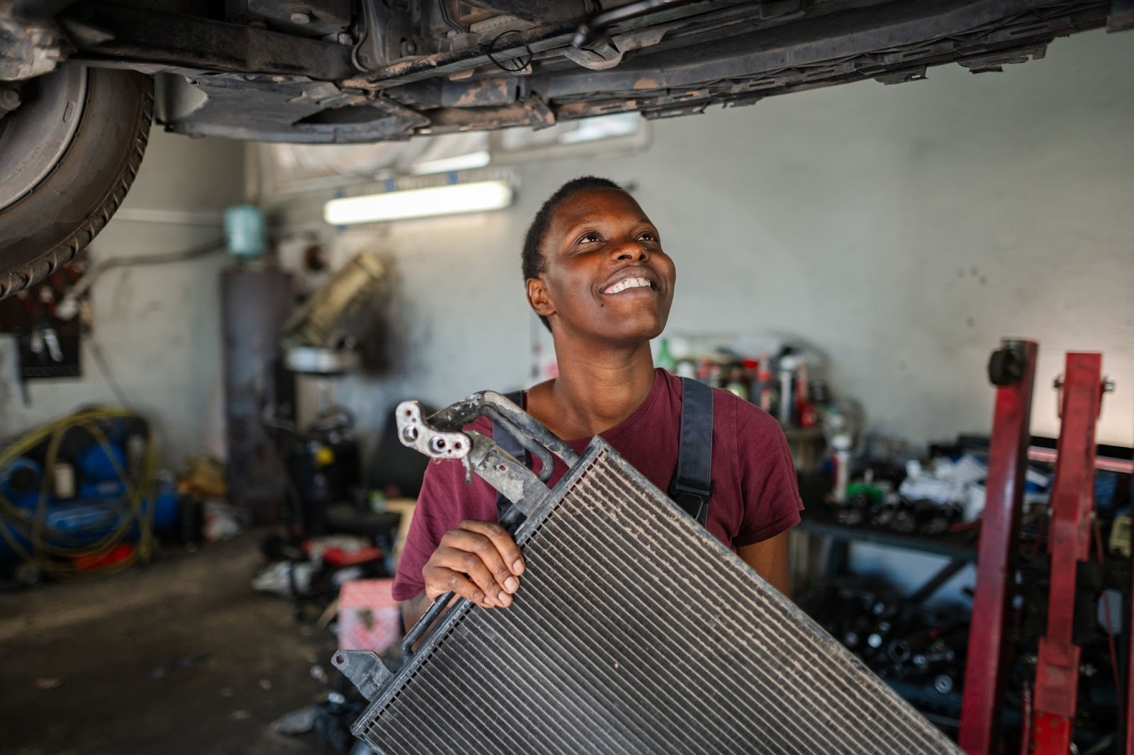 Female auto mechanic holding a radiator while inspecting the car underneath in a professional repair shop