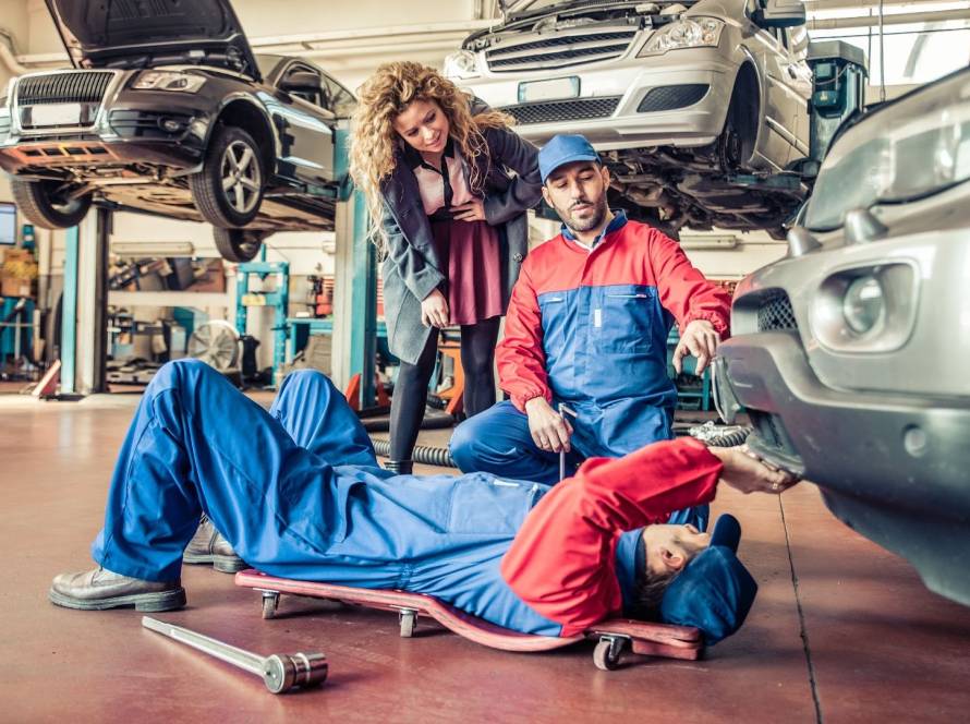 Two mechanics repair a damaged car as the concerned owner watches, with other vehicles lifted in the background