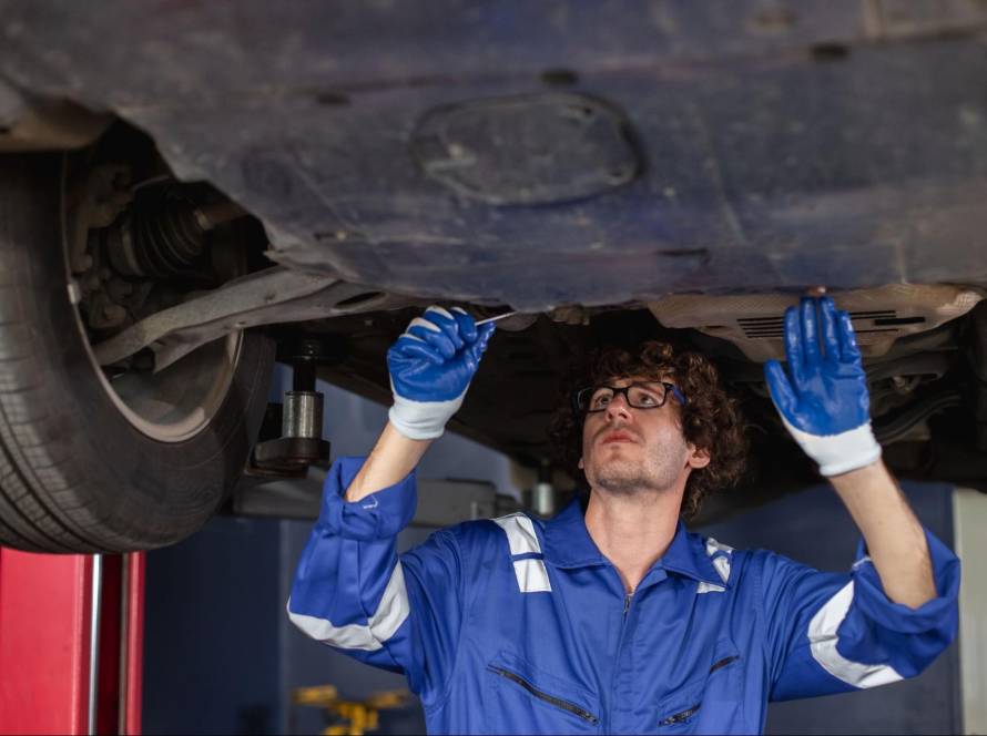 Auto mechanic wearing eyeglasses and a blue coverall, inspecting a vehicle’s muffler and suspension.