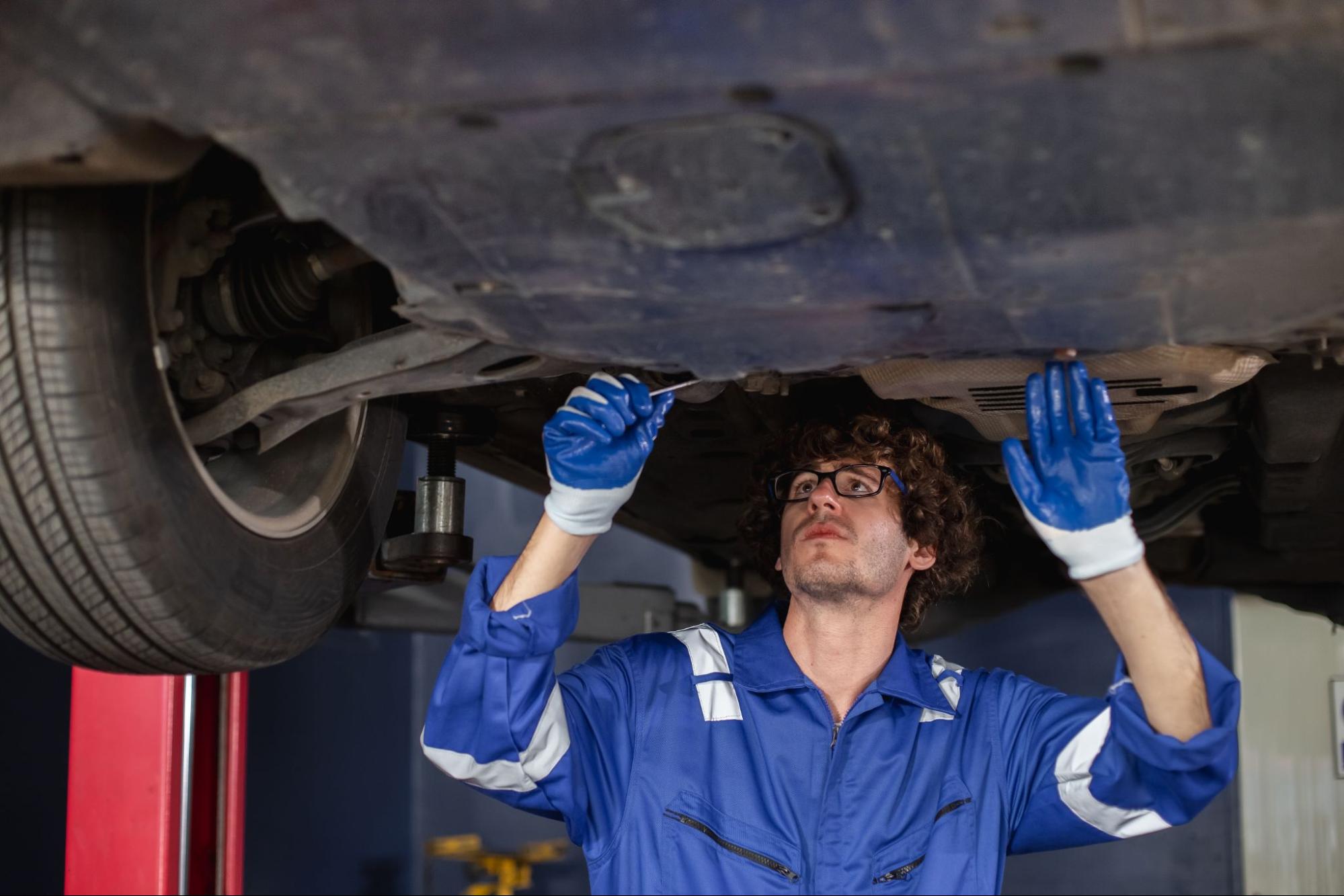 Auto mechanic wearing eyeglasses and a blue coverall, inspecting a vehicle’s muffler and suspension.